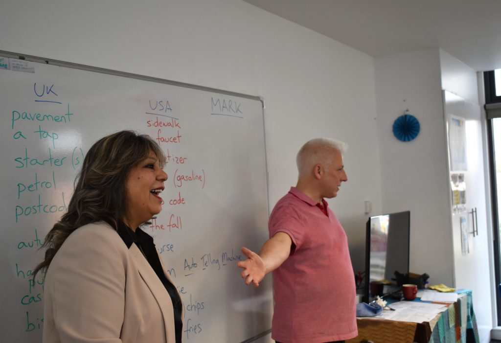 "Close-up of a woman in a beige blazer and a man in a pink shirt standing in front of a whiteboard. The woman is smiling, and the man gestures as if explaining something. The whiteboard displays comparisons between UK and USA English words written in blue and red markers."