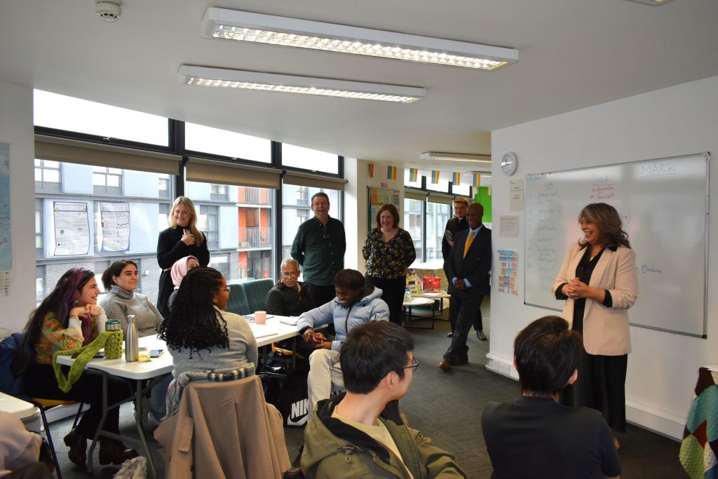 "Group of people in a classroom setting with a diverse group of attendees seated at tables, listening to a woman in a beige blazer speaking at the front. Several others stand at the back of the room, smiling and observing. The whiteboard displays a list of UK and USA English word comparisons, and colourful flags decorate the space."