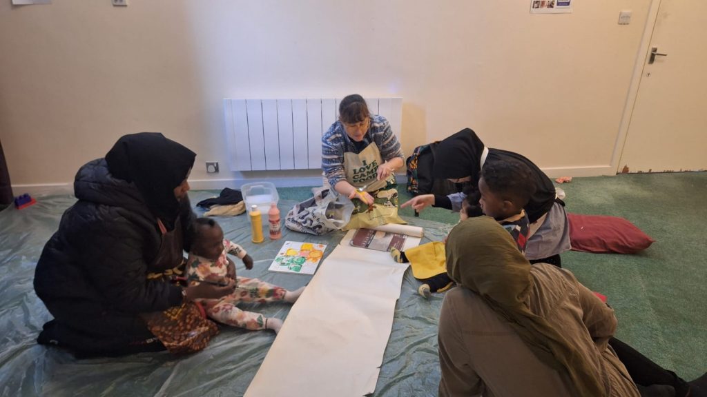 Group of mothers and toddlers engaging in a creative activity together, with a facilitator demonstrating painting techniques. The setting is a relaxed and welcoming indoor space with art supplies and a laid-back atmosphere.