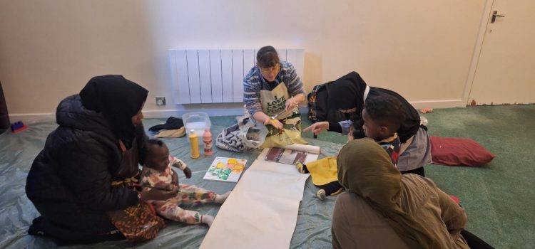 Group of mothers and toddlers engaging in a creative activity together, with a facilitator demonstrating painting techniques. The setting is a relaxed and welcoming indoor space with art supplies and a laid-back atmosphere.